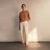 View 1 of product A woman stands barefoot indoors against a neutral wall, wearing Turnbull & Asser Women's Cream Cashmere Knitted Tabitha Bottoms and a light brown sweater. Soft sunlight casts a gentle shadow on the wall beside her.
