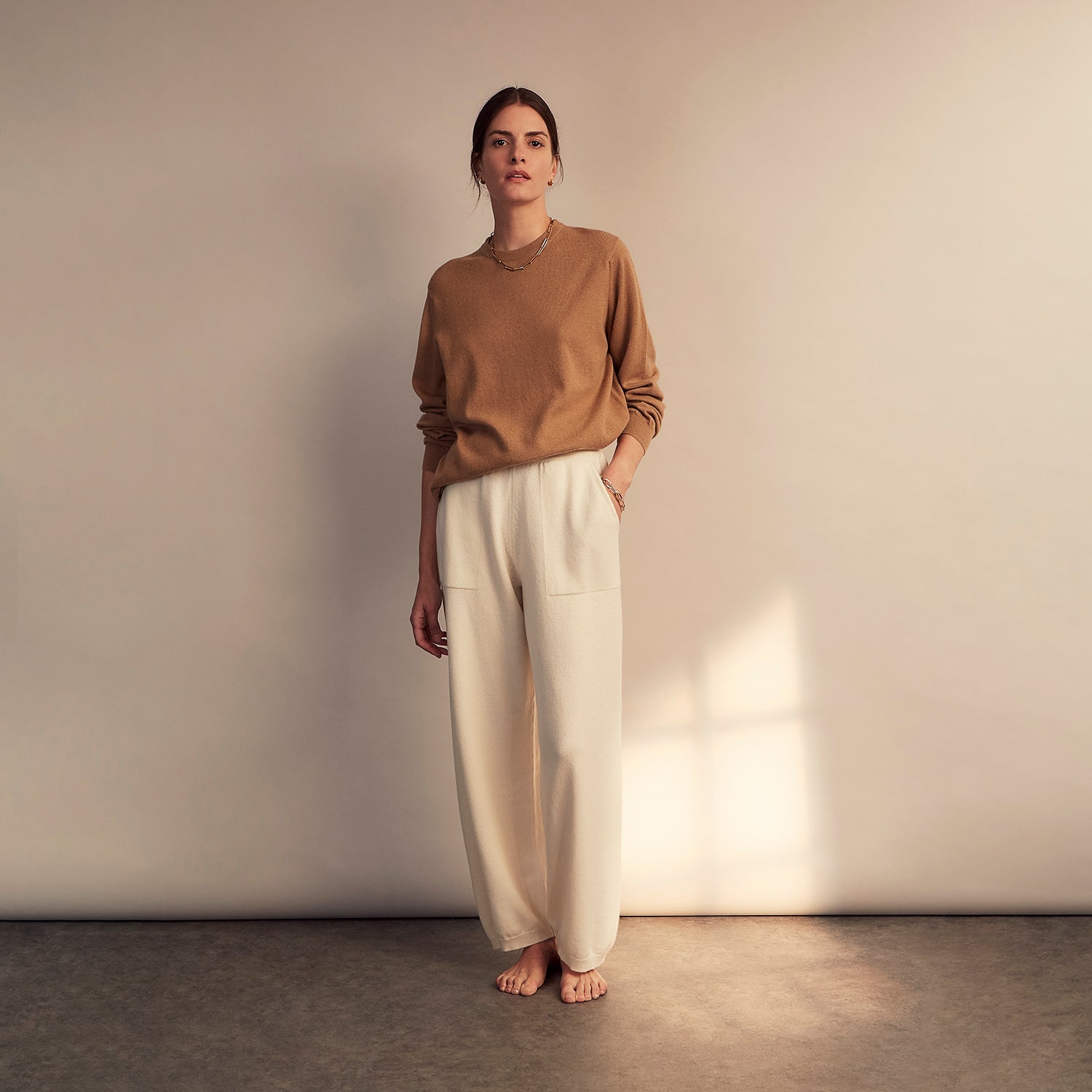 View 1 of product A woman stands barefoot indoors against a neutral wall, wearing Turnbull & Asser Women's Cream Cashmere Knitted Tabitha Bottoms and a light brown sweater. Soft sunlight casts a gentle shadow on the wall beside her.