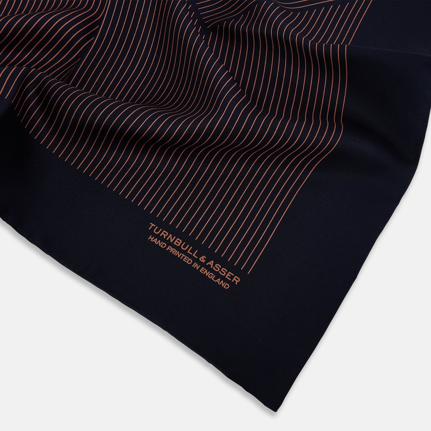 View 2 of product The Turnbull & Asser Navy and Blush Stripe Silk Pocket Square features thin blush stripes on navy silk, finished with "TURNBULL & ASSER HAND PRINTED IN ENGLAND" in the corner, highlighting its refined English craftsmanship.