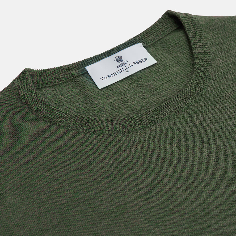 A man stands facing the camera against a plain light background, wearing the Turnbull & Asser Bottle Green Merino Kingston Crew Neck Jumper with dark pants and black shoes. His arms rest at his sides and he has a neutral expression.