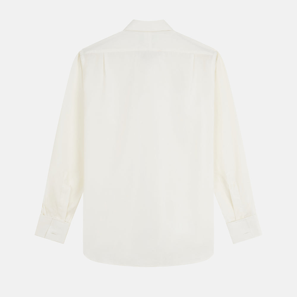 A man in a Cream Silk Regular Fit Ruffle-Front Wilfred Shirt by Turnbull & Asser stands against a dark background, one hand on a chair, gazing thoughtfully to the side.