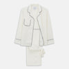 View 1 of product The Women's Harriet Cream Silk Pyjama Set by Turnbull & Asser features a white sand-washed silk button-up top with black piping and chest pocket, plus matching drawstring pants, all shown against a white background.