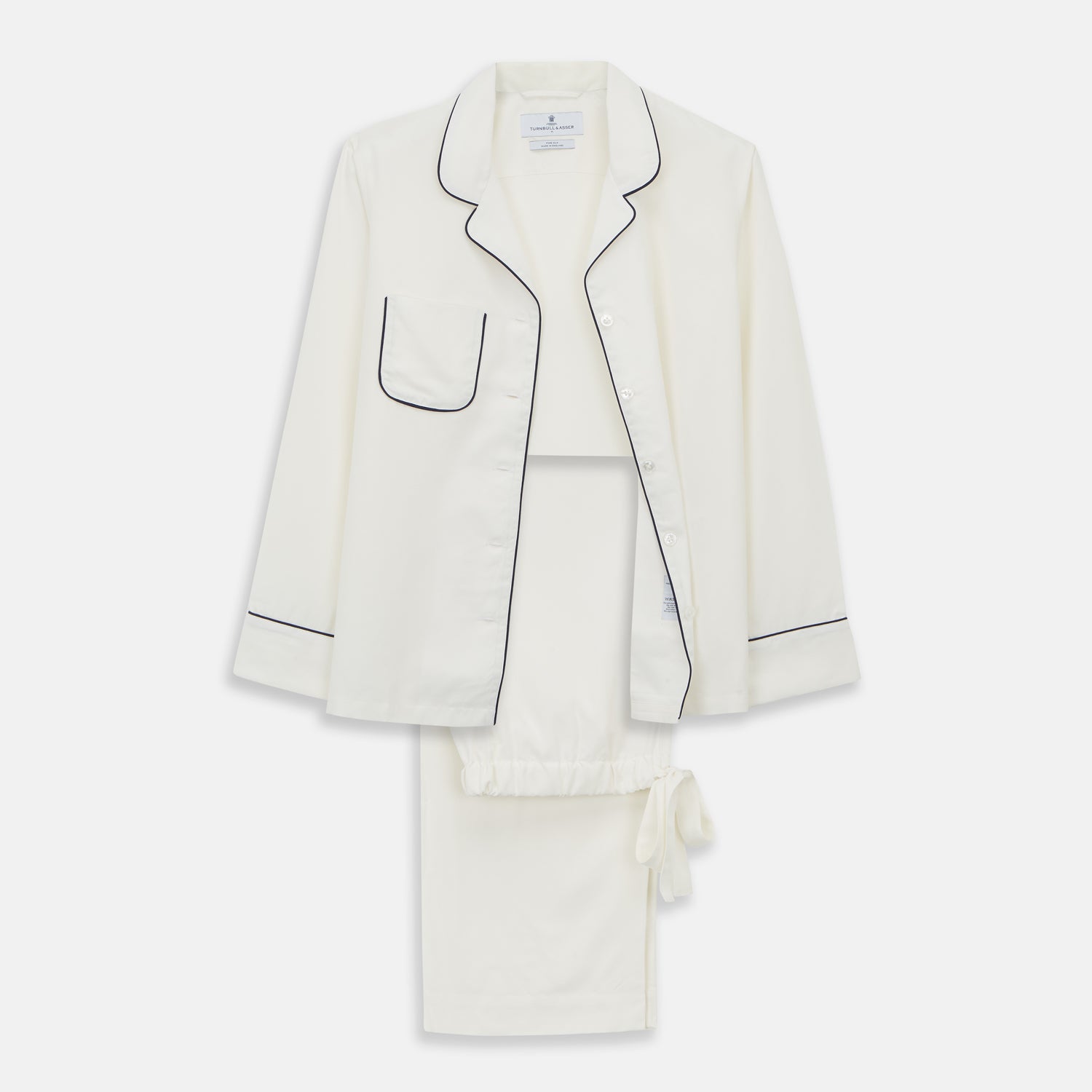 View 1 of product The Women's Harriet Cream Silk Pyjama Set by Turnbull & Asser features a white sand-washed silk button-up top with black piping and chest pocket, plus matching drawstring pants, all shown against a white background.