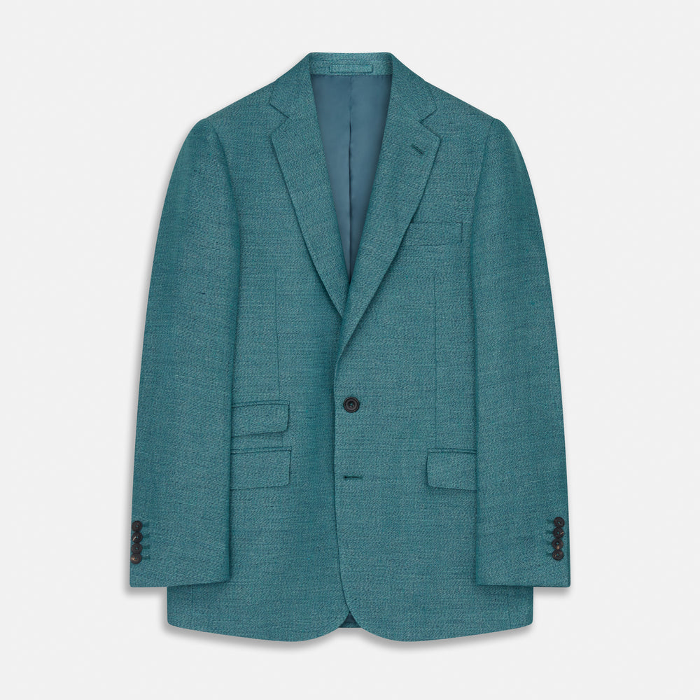 A man with short brown hair and a neutral expression stands against a plain background, wearing a Teal Linen Barrington Blazer by Turnbull & Asser, a light pink dress shirt, white trousers, brown loafers, and a patterned pocket square.