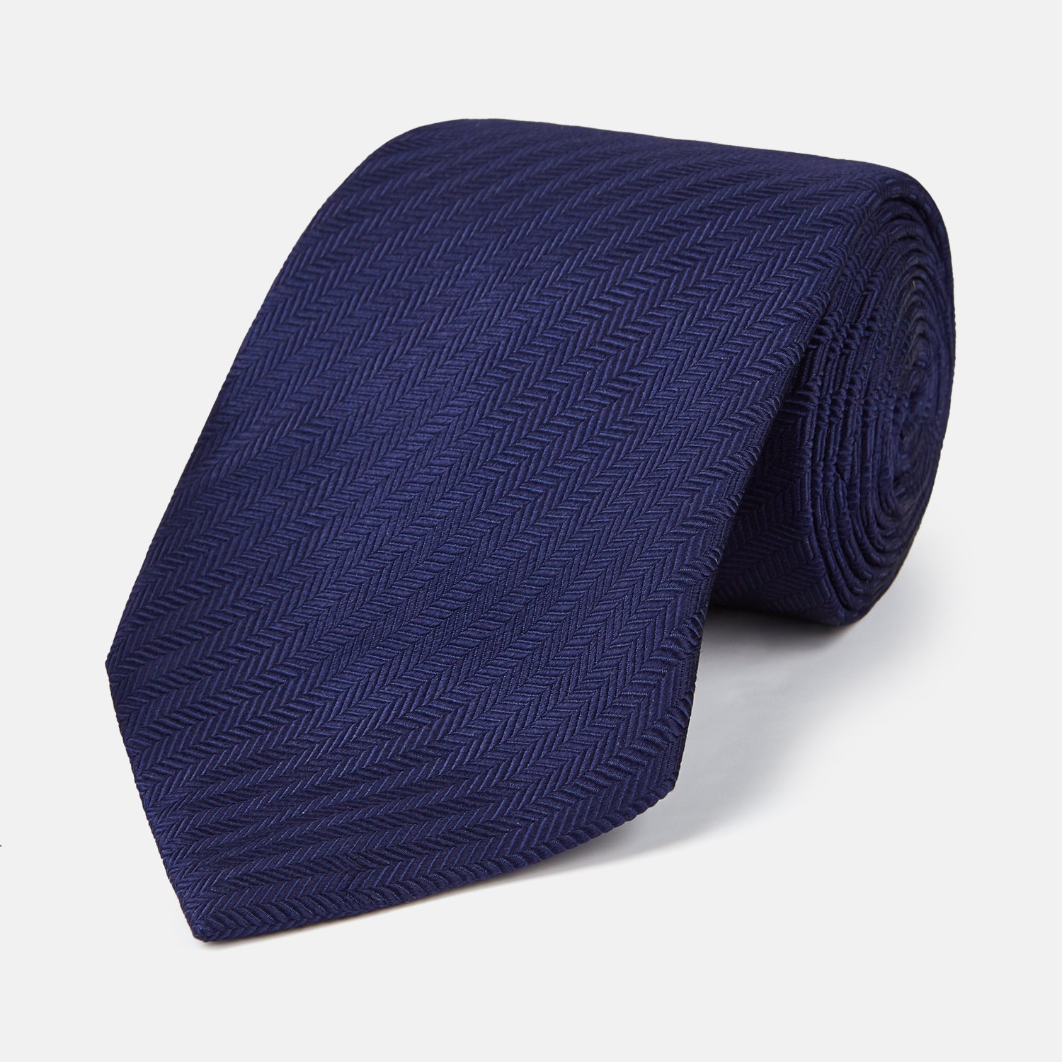 View 1 of product The PLAIN HERRINGBONE NAVY TIE by Turnbull & Asser, crafted from silk with a subtle pattern, is elegantly rolled and displayed against a plain white background.