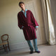 A person stands indoors by a window, wearing the Turnbull & Asser Burgundy Cotton-Cashmere Kimono Gown over striped pajamas, with matching slippers, next to cream curtains and a wooden chair against a pale wall.