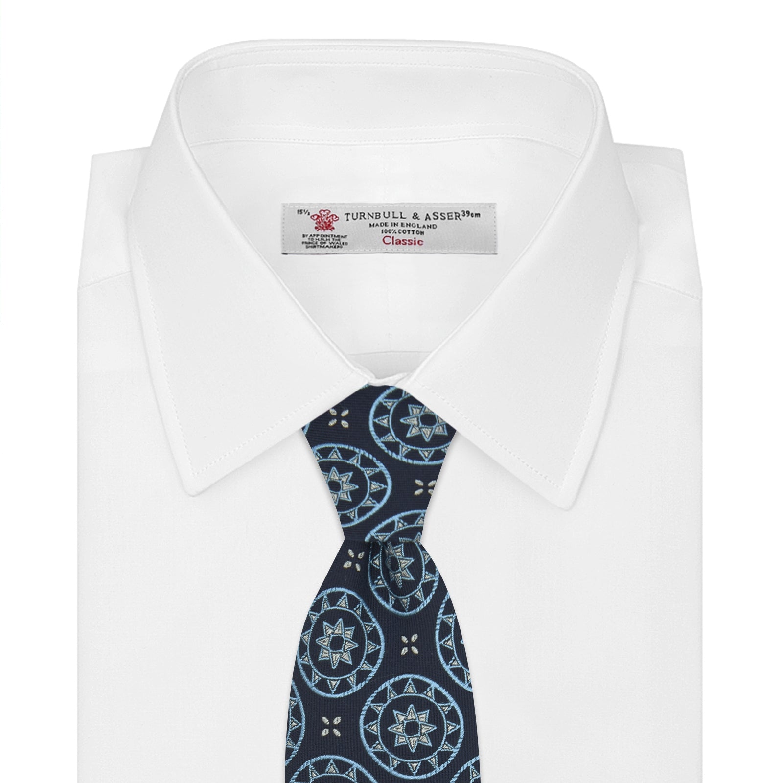 View 2 of product The Jacquard Silk Geometric Tie by Turnbull & Asser features a navy blue base with light blue and white star and circle motifs, shown rolled on a white background.
