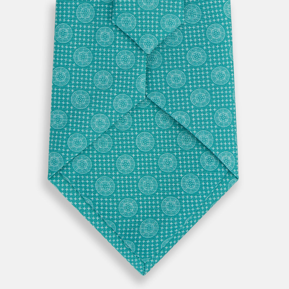 The Turnbull & Asser Turquoise Medallion Silk Tie features light blue and white circular motifs on fine English silk—an elegant men’s accessory, rolled up and displayed on a white background.