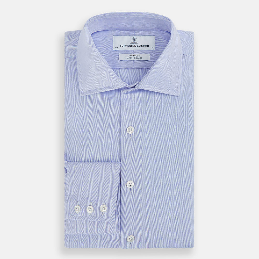 The Turnbull & Asser BLUE HERRINGBONE TAILORED FIT SHIRT WITH KENT COLLAR features long sleeves and buttoned cuffs, offering a timeless formal look in a classic light blue herringbone fabric, laid flat on a white background.