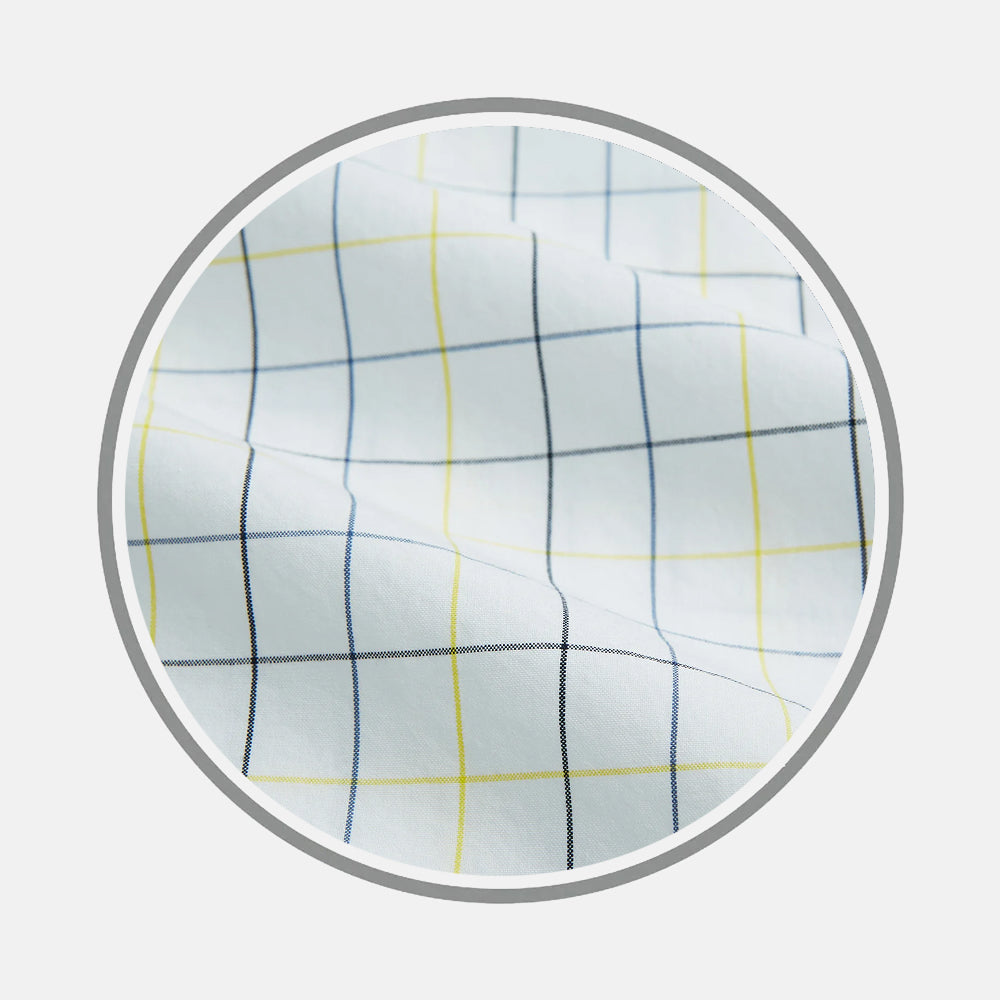 A close-up of Turnbull & Asser's Yellow and Blue Grid Check Fabric shows its white base with thin blue and yellow lines in a grid pattern, slightly wrinkled and displayed within a circular frame on a plain gray background.