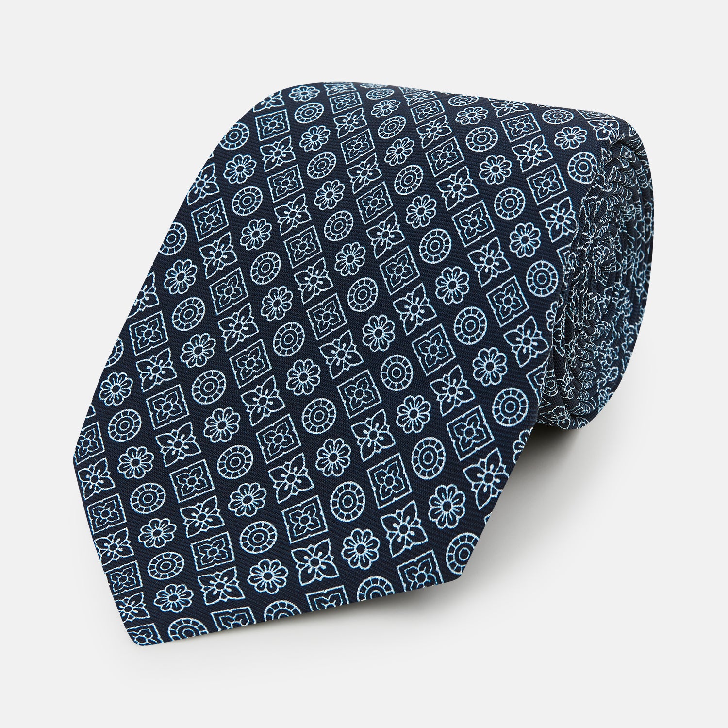 View 1 of product The Turnbull & Asser Navy Floral Tile Pattern Printed Silk Tie features light blue geometric and floral motifs in a diamond grid, elegantly displayed and crafted from fine English silk.
