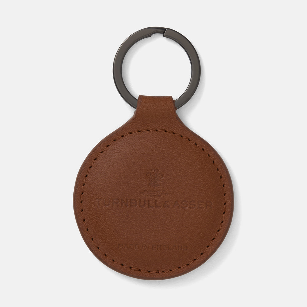 A tan circular leather fob holder by Turnbull & Asser, featuring visible stitching, embossed branding, and 'Made in England,' attached to a metal key ring.