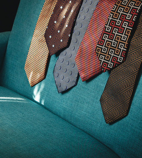 Five patterned neckties in various colors and geometric designs are draped over the arm of a teal fabric chair, with sunlight casting shadows on the seat.