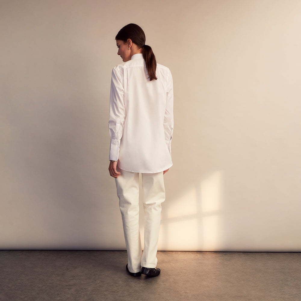 A person with curly hair wears the Turnbull and The Deck White Cotton Minimalist Shirt by Turnbull & Asser, paired with pants. Posed against a plain background, their minimalist silhouette is highlighted in this black and white image.