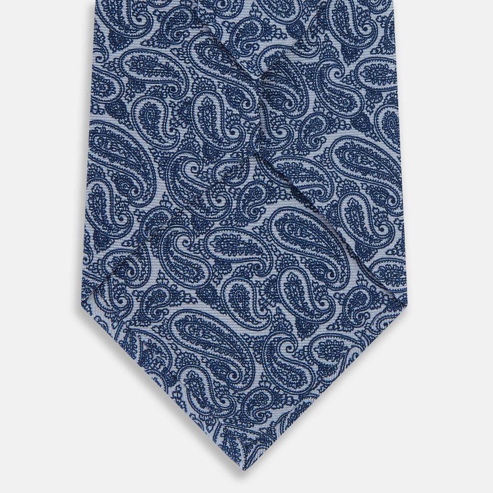 The Turnbull & Asser Pale Blue Paisley Silk Tie, featuring a blue paisley pattern on a pale blue background, is shown rolled up on a plain white surface.