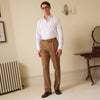 View 1 of product A man in glasses, a white dress shirt, Turnbull & Asser Tan Wool Pleated Jasper Trousers, and dark brown shoes stands in a light room with a radiator, antique desk, round mirror, and wall sconce.