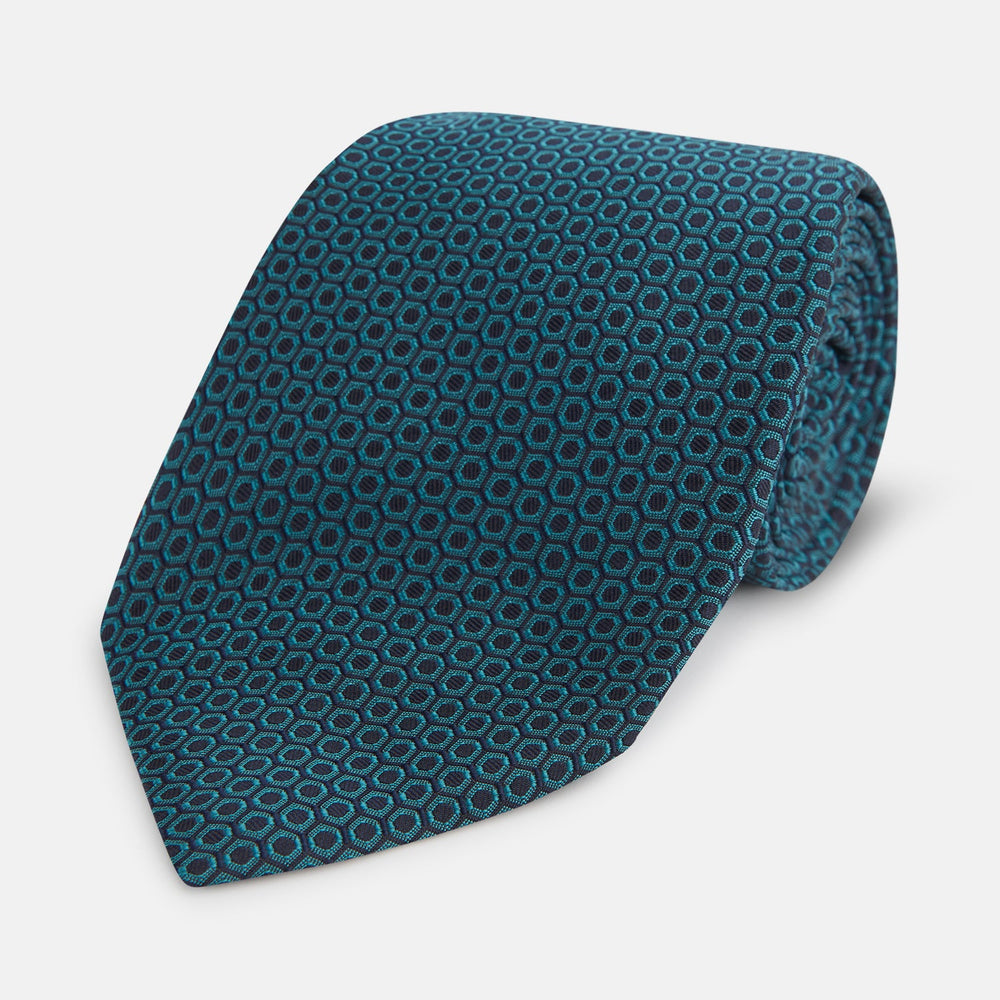 The Turnbull & Asser Turquoise Micro Hexagon Tie features refined British elegance with its small blue hexagonal pattern on a lighter background, neatly rolled and displayed on a plain white surface.