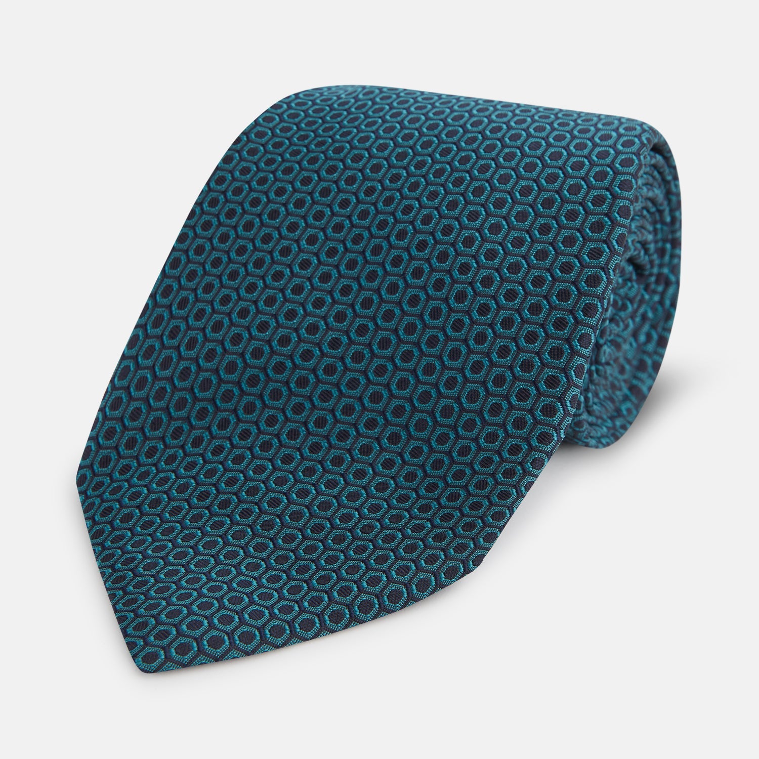 View 1 of product The Turnbull & Asser Turquoise Micro Hexagon Tie features refined British elegance with its small blue hexagonal pattern on a lighter background, neatly rolled and displayed on a plain white surface.