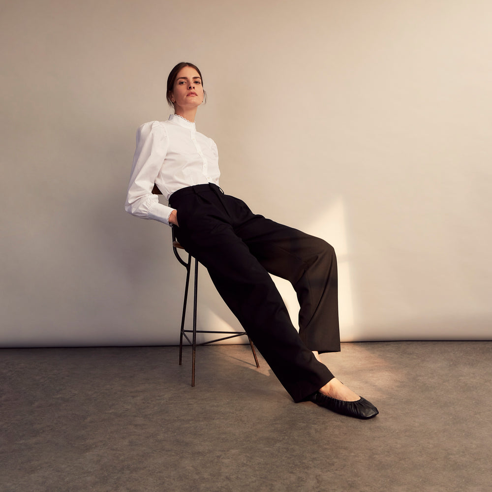 A woman stands in profile, looking down with hands in her pockets, wearing the Turnbull & Asser Turnbull and The Deck White Cotton Expressionist Shirt, paired with high-waisted black-and-white houndstooth pants.