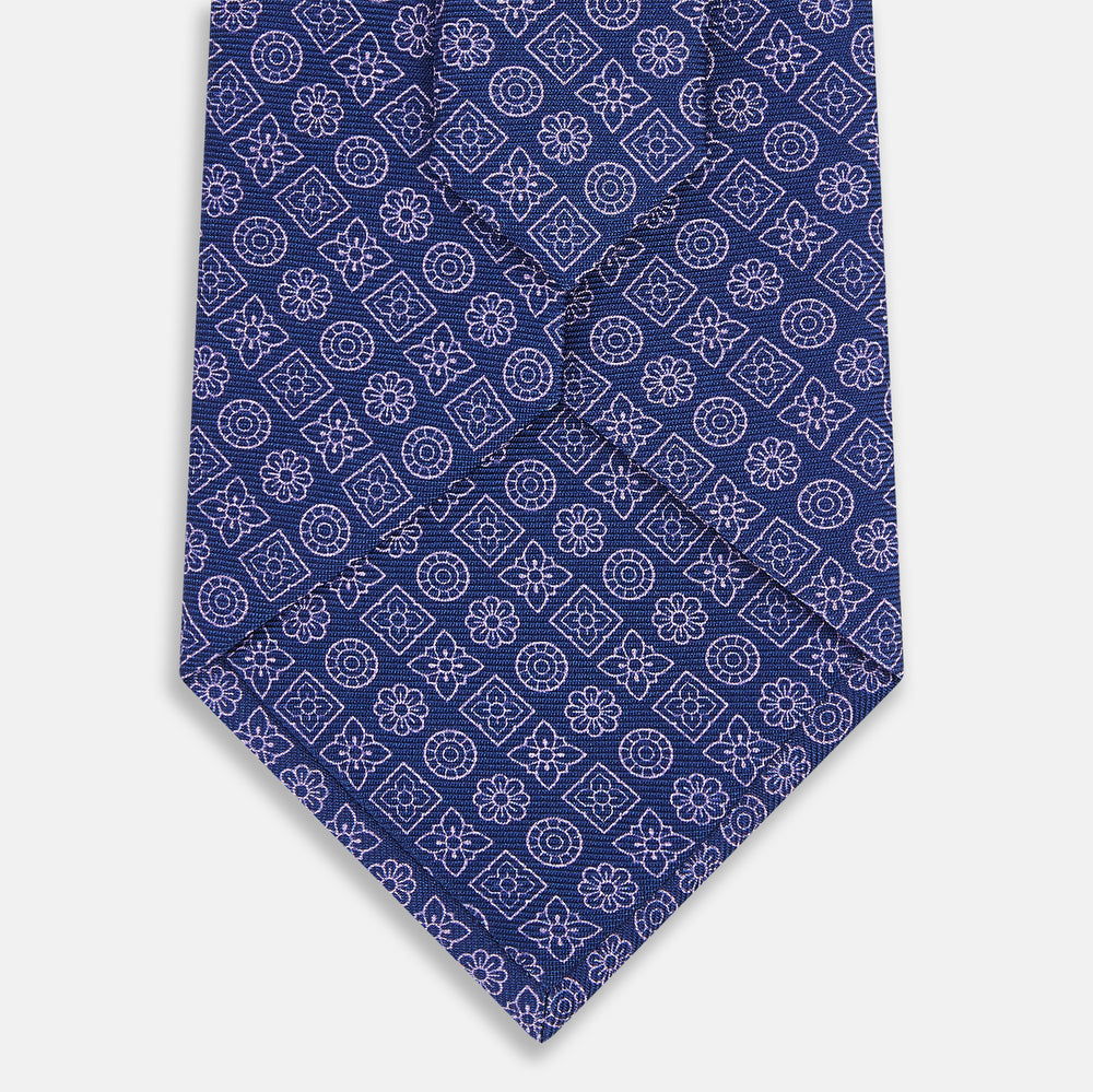 The Turnbull & Asser Azzurro Floral Tile Pattern Printed Silk Tie features blue silk with a geometric design of white circles, squares, and floral motifs, displayed on a plain white background.