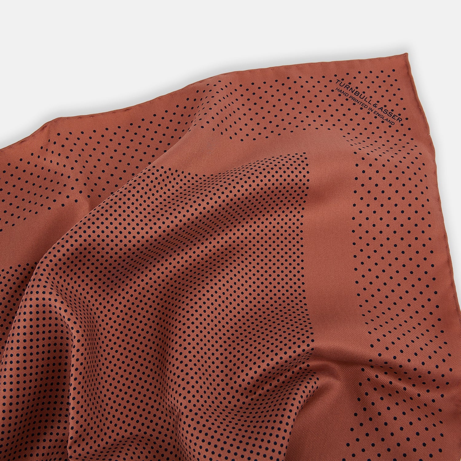 View 2 of product The Turnbull & Asser Blush Dot Silk Pocket Square features a rust hue with black polka dots, a bordered design, and hand-rolled edges, shown folded against a white background.