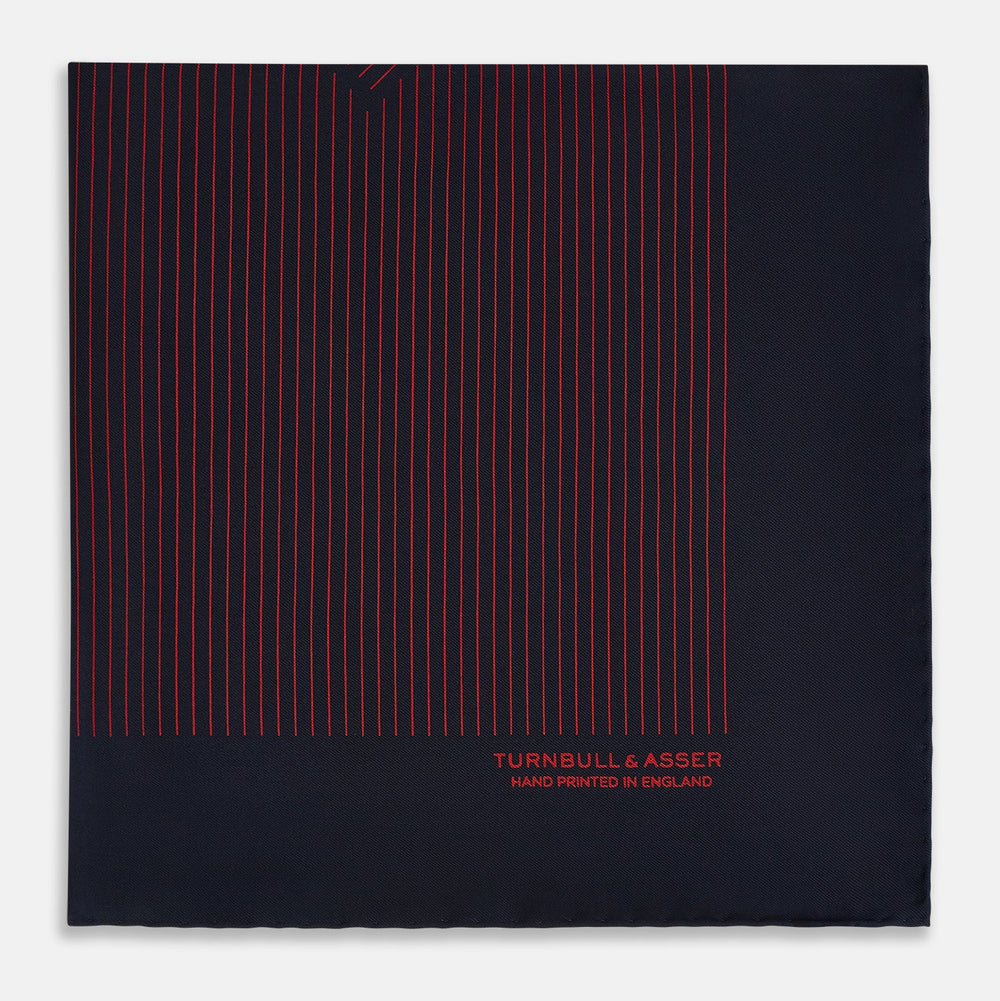 The Turnbull & Asser Navy and Red Stripe Silk Pocket Square features thin red stripes on navy silk, with "HAND PRINTED IN ENGLAND" in red at the corner. A folded corner shows its hand-rolled edges and refined design.