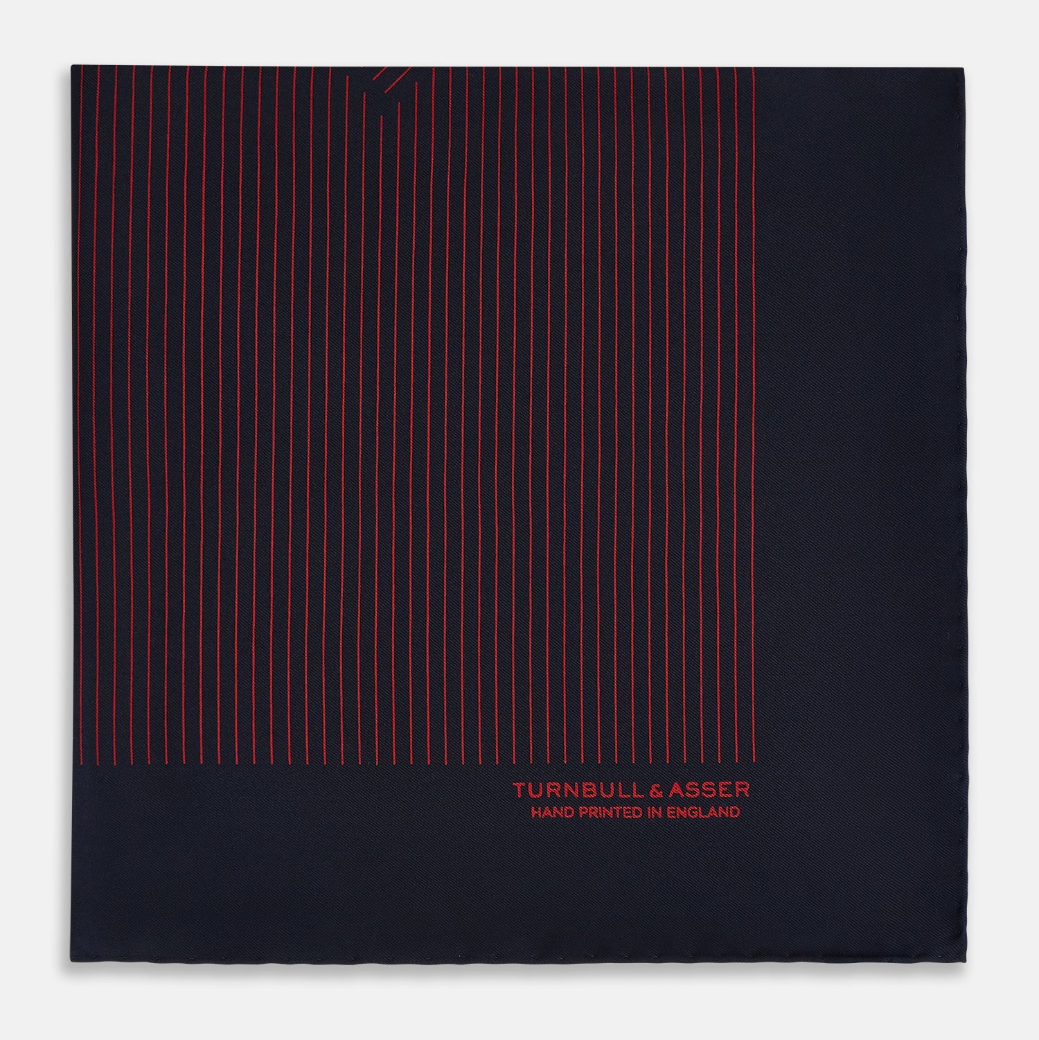 View 3 of product The Turnbull & Asser Navy and Red Stripe Silk Pocket Square features thin red stripes on navy silk, with "HAND PRINTED IN ENGLAND" in red at the corner. A folded corner shows its hand-rolled edges and refined design.