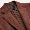 View 3 of product A man stands in a lush garden, hands in pockets and relaxed, wearing the Turnbull & Asser Red Lambswool Herringbone Bern Blazer, a striped dress shirt, patterned scarf, and dark trousers.