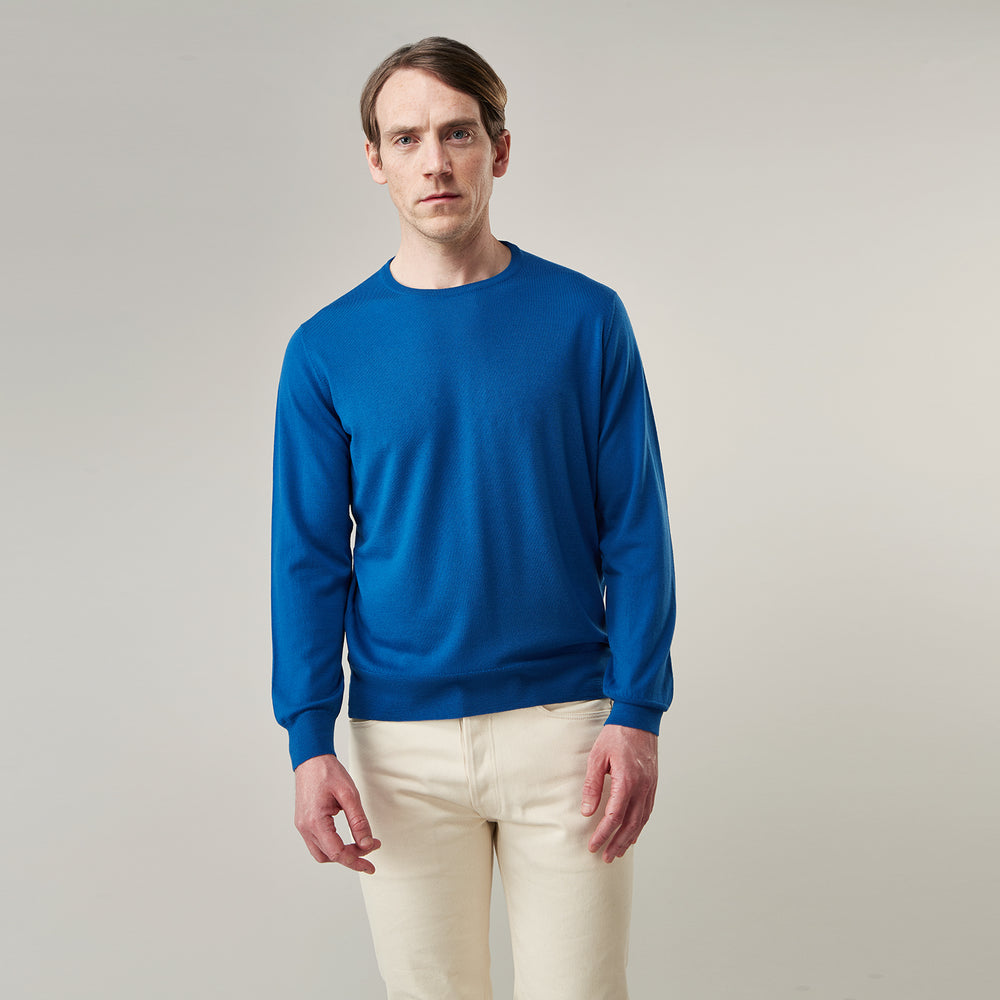 A man with light skin and short brown hair stands against a plain background, wearing the Turnbull & Asser Ocean Blue Merino Kingston Crew Neck Jumper and cream-colored pants, looking directly at the camera with a neutral expression.