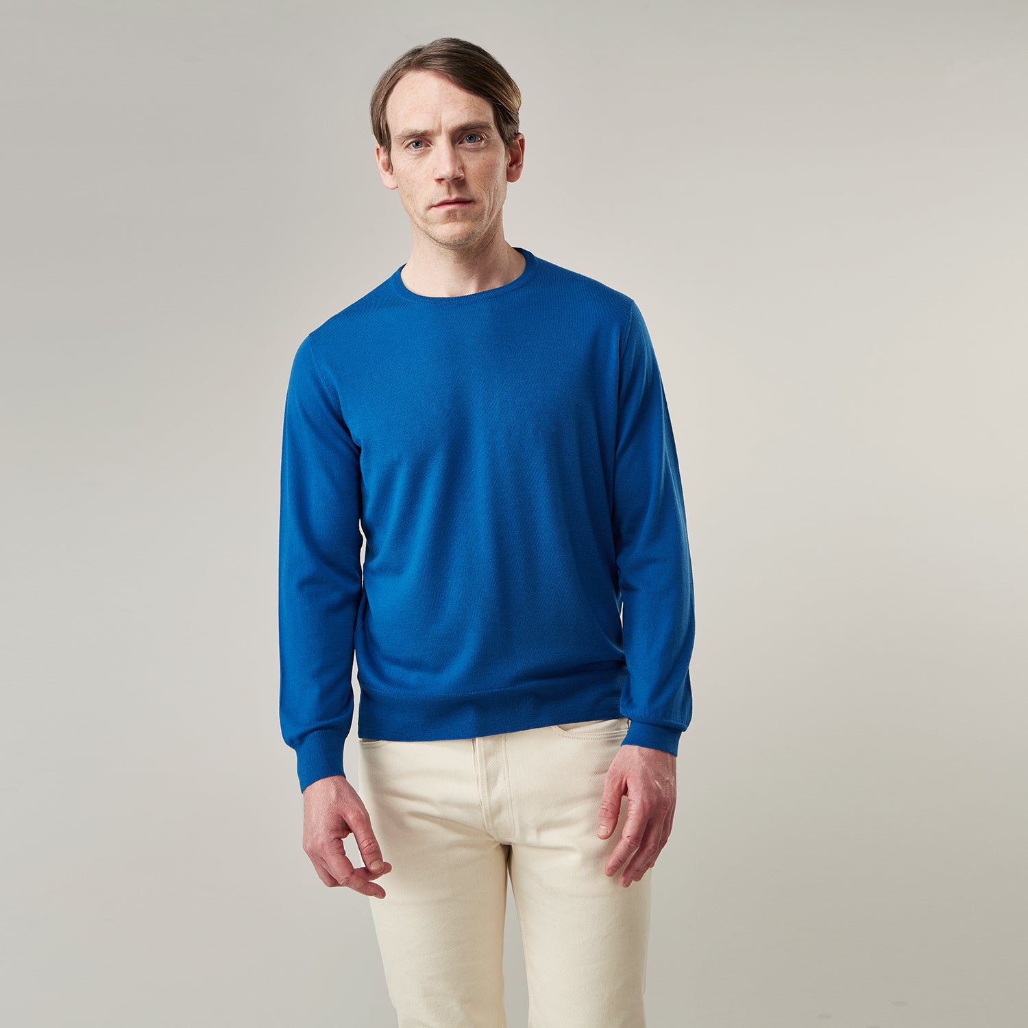 View 1 of product A man with light skin and short brown hair stands against a plain background, wearing the Turnbull & Asser Ocean Blue Merino Kingston Crew Neck Jumper and cream-colored pants, looking directly at the camera with a neutral expression.