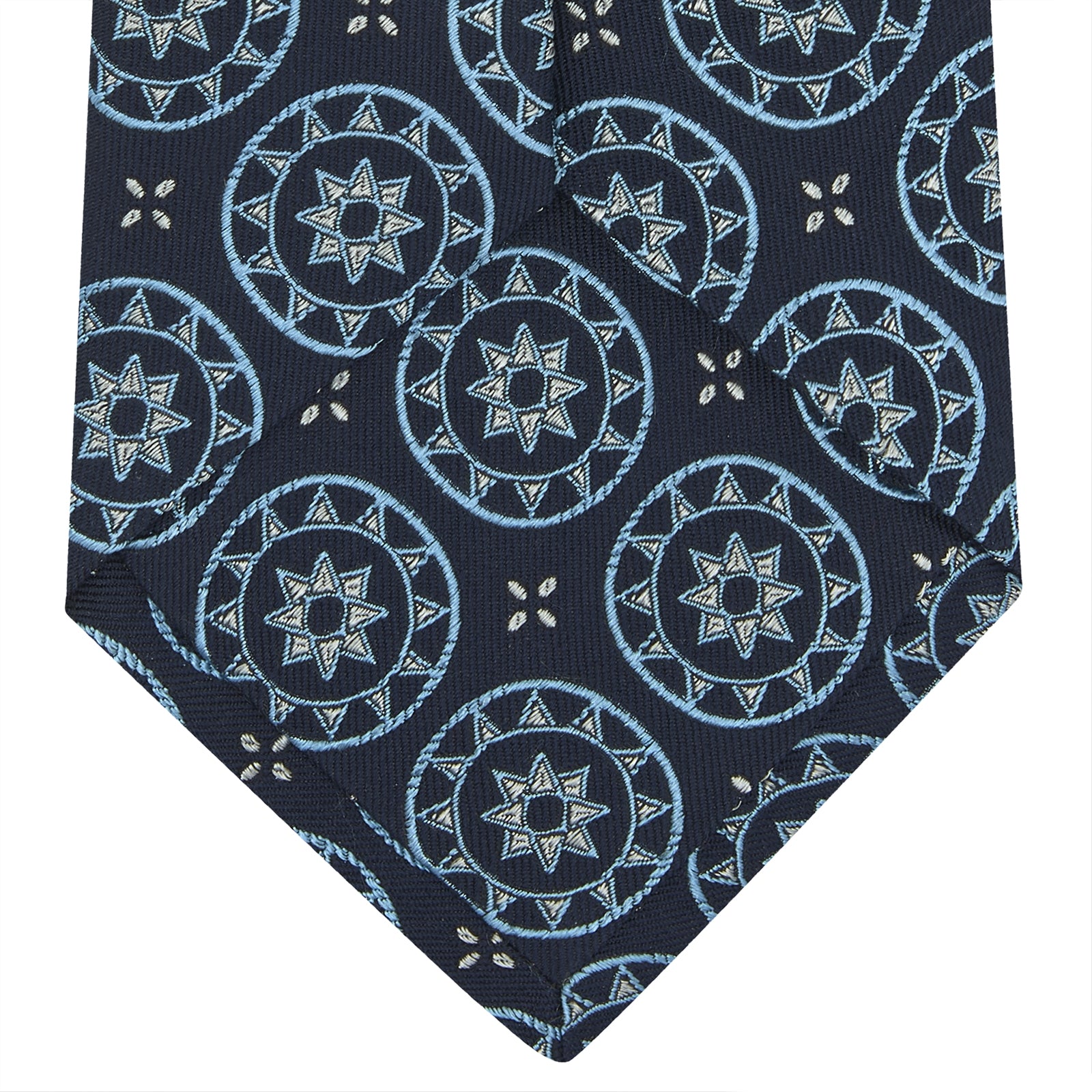 View 3 of product The Jacquard Silk Geometric Tie by Turnbull & Asser features a navy blue base with light blue and white star and circle motifs, shown rolled on a white background.