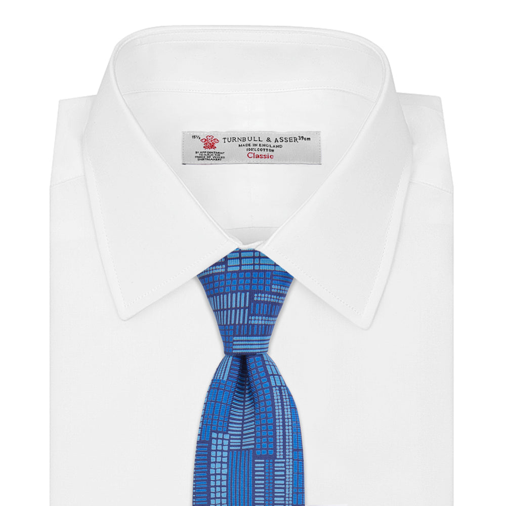 The Turnbull & Asser Blue Motif Printed Foulard Silk Tie showcases a geometric pattern in varying blue shades, making it a striking and essential accessory for any wardrobe.