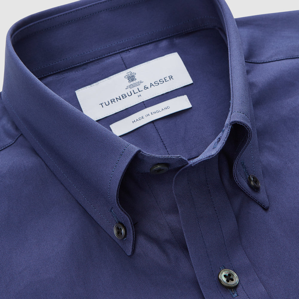 The Blue Piece Dyed Weekend Fit Suffolk Shirt by Turnbull & Asser features a button-down Dorset collar, chest pocket, white buttons, and long sleeves in classic navy, laid flat against a white background.