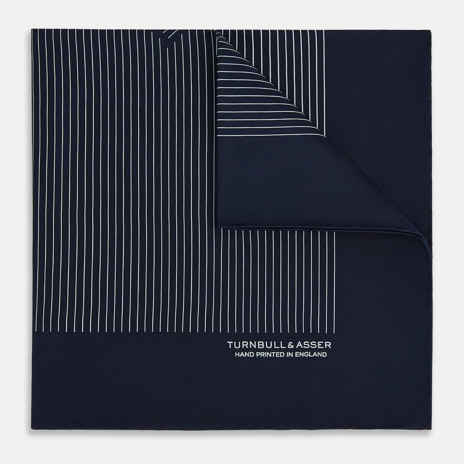 View 1 of product The Navy and White Stripe Silk Pocket Square by Turnbull & Asser features thin white vertical stripes and "TURNBULL & ASSER HAND PRINTED IN ENGLAND" in one corner, with a folded edge showing the reverse of this elegant silk accessory.