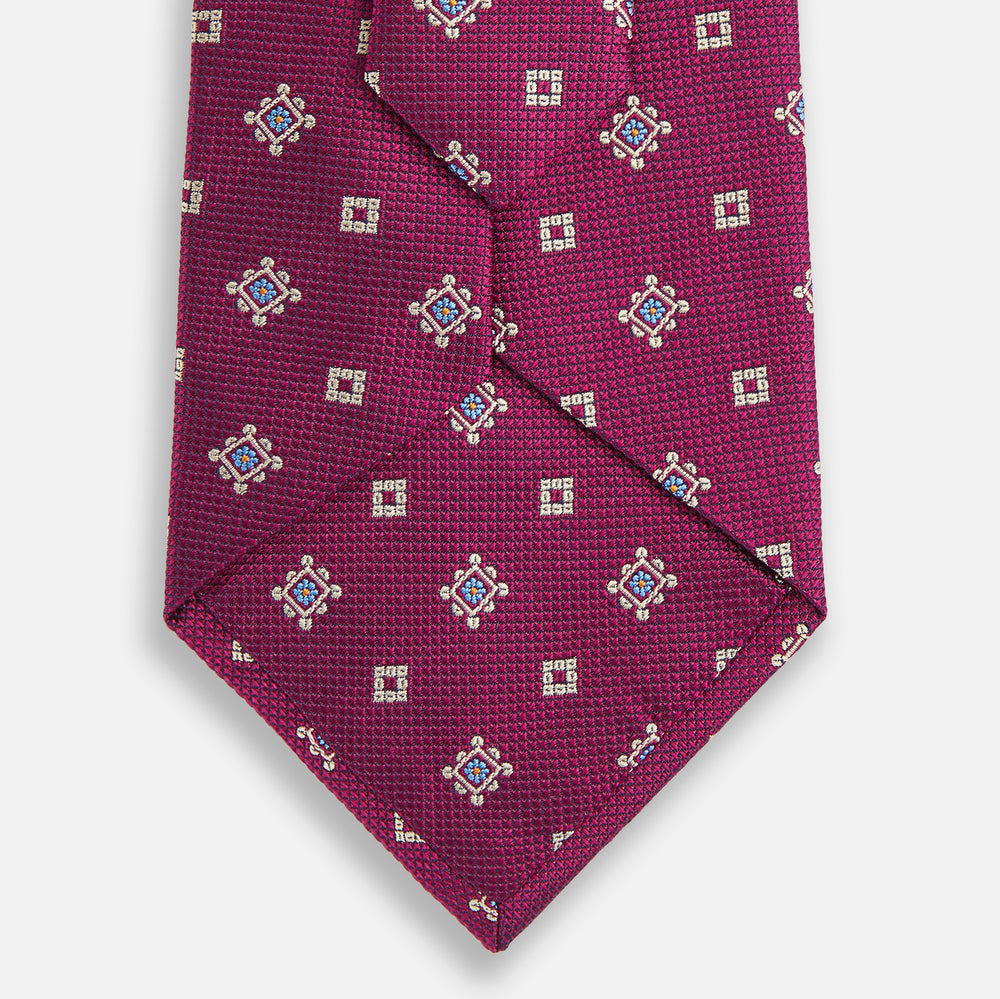 The Turnbull & Asser Purple Motif Silk Tie, rolled up and displayed on a light gray background, features small blue and white geometric designs—an elegant British touch for any formal event.