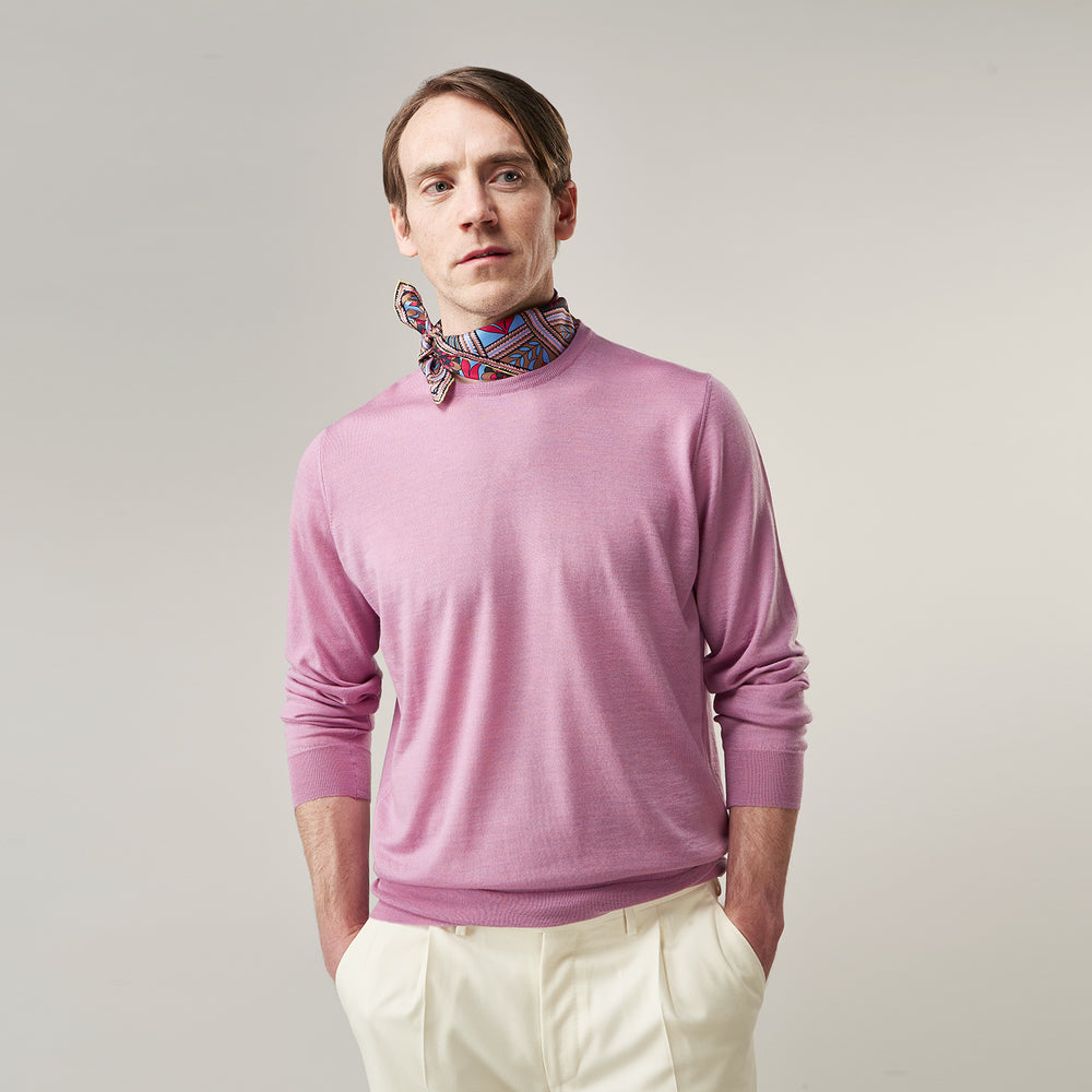 Wearing Turnbull & Asser’s Pink Merino Kingston Crew Neck Jumper, white pants, and a colorful scarf, a person stands against a plain light gray background with hands in their pockets.