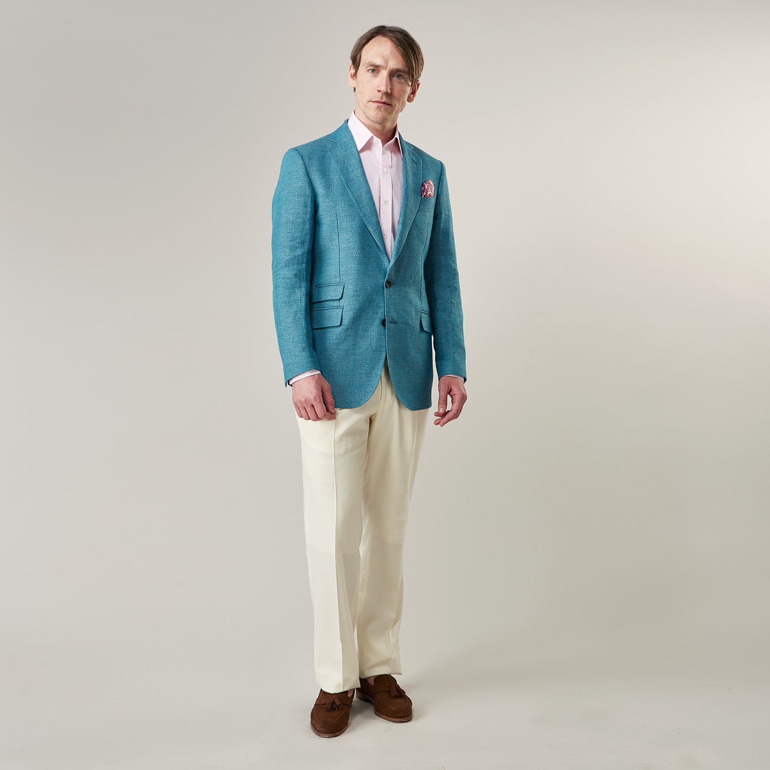 View 5 of product A man with short brown hair and a neutral expression stands against a plain background, wearing a Teal Linen Barrington Blazer by Turnbull & Asser, a light pink dress shirt, white trousers, brown loafers, and a patterned pocket square.