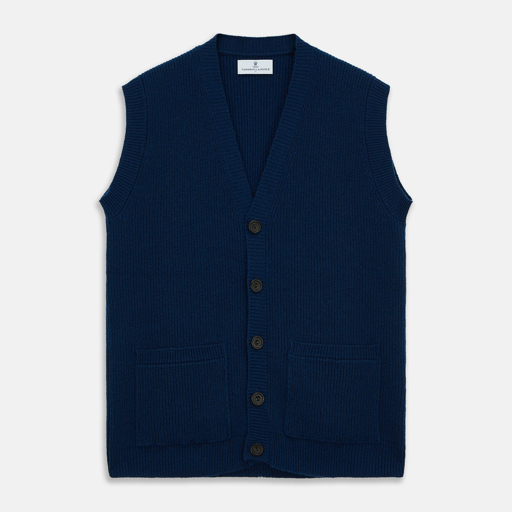 A man with a shaved head wears a Turnbull & Asser Navy Sleeveless Cashmere Cardigan over a white dress shirt with rolled-up sleeves and dark trousers, standing against a plain light gray background.
