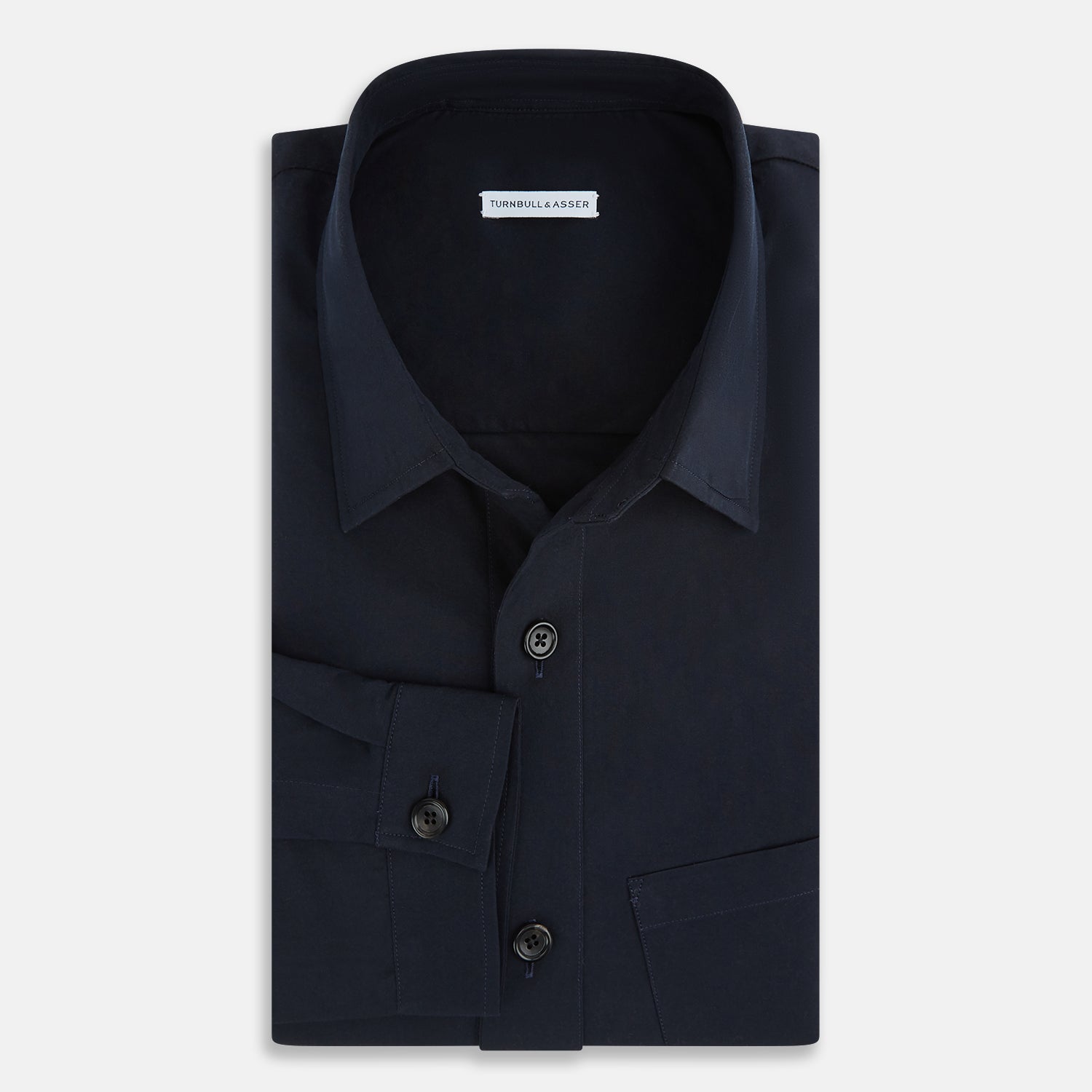 View 5 of product The Midnight Blue Hyde Overshirt by Turnbull & Asser is a plain, long-sleeve shirt in dark navy sanforised cotton with a single chest pocket, spread collar, and black buttons, shown laid flat on a white background.