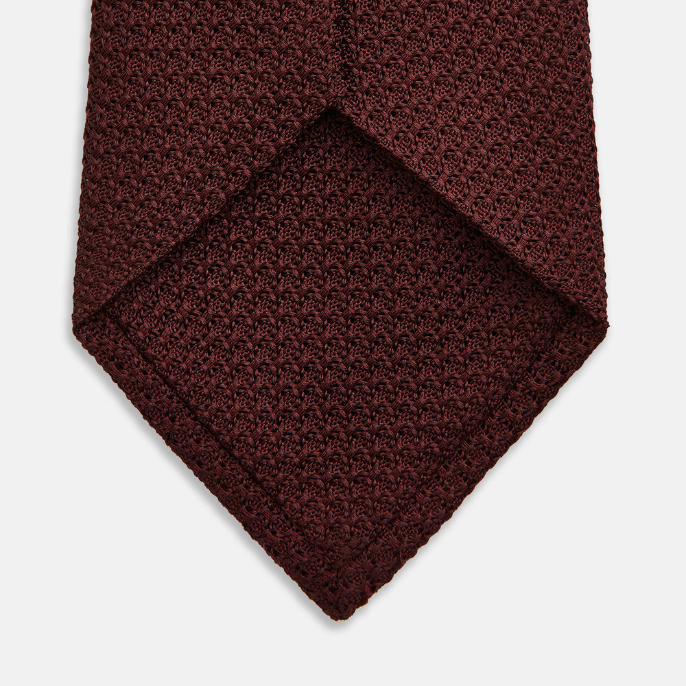 The Turnbull & Asser Ruby Red Knitted Tie, shown rolled-up against a white background, is a textured silk accessory that adds versatile sophistication to any gentleman's wardrobe.