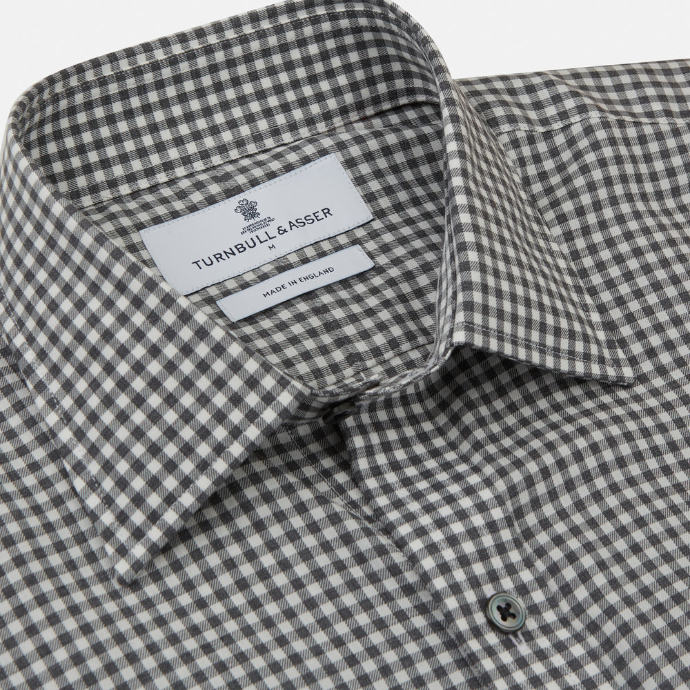 A person with a shaved head models the Turnbull & Asser Grey Check Weekend Fit Flannel Shirt with a Derby collar and 1-button cuffs, sleeves rolled up, paired with dark trousers against a plain background, gazing directly at the camera.