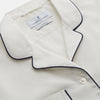 View 3 of product The Women's Harriet Cream Silk Pyjama Set by Turnbull & Asser features a white sand-washed silk button-up top with black piping and chest pocket, plus matching drawstring pants, all shown against a white background.