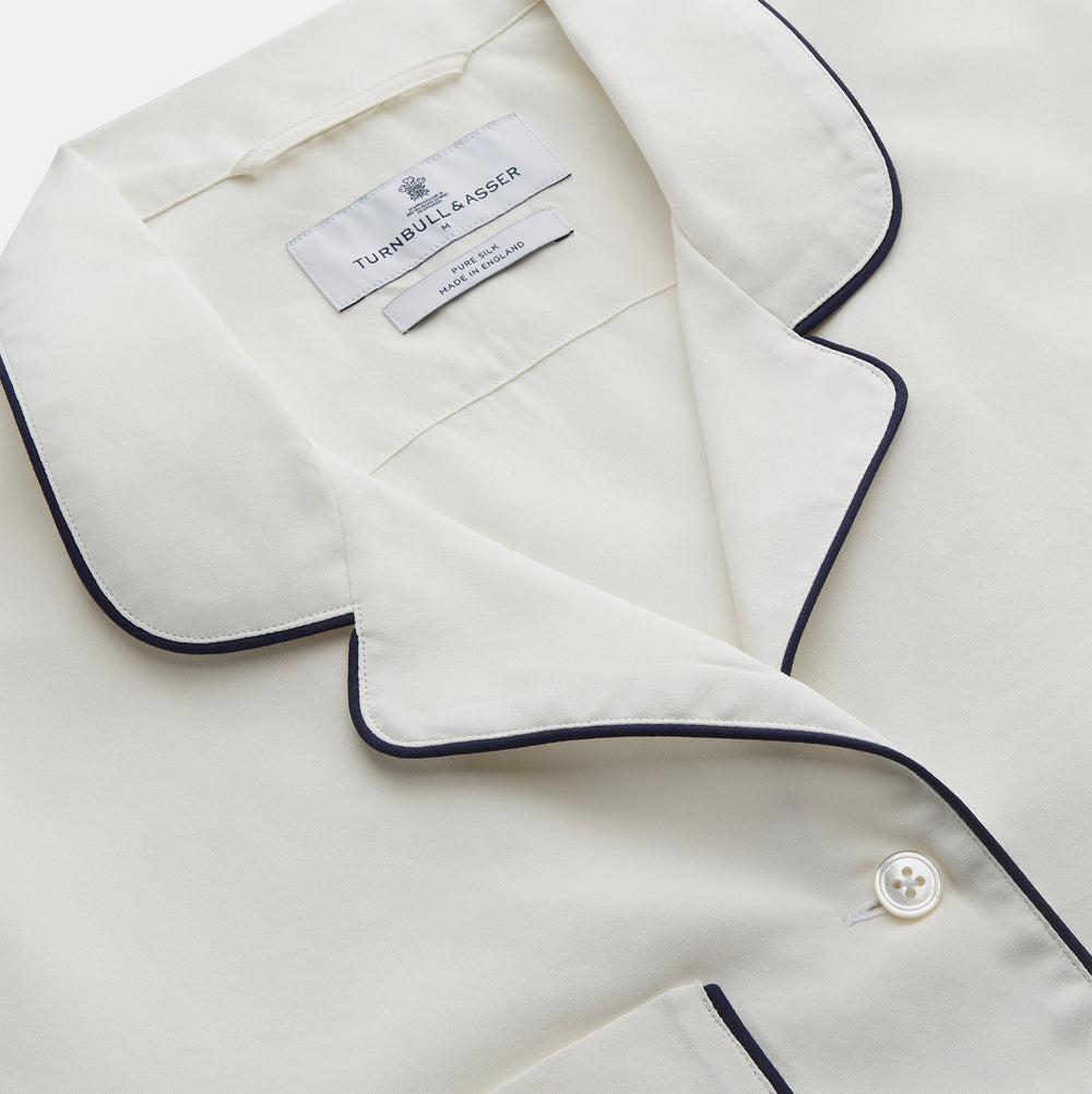 The Women's Harriet Cream Silk Pyjama Set by Turnbull & Asser features a white sand-washed silk button-up top with black piping and chest pocket, plus matching drawstring pants, all shown against a white background.