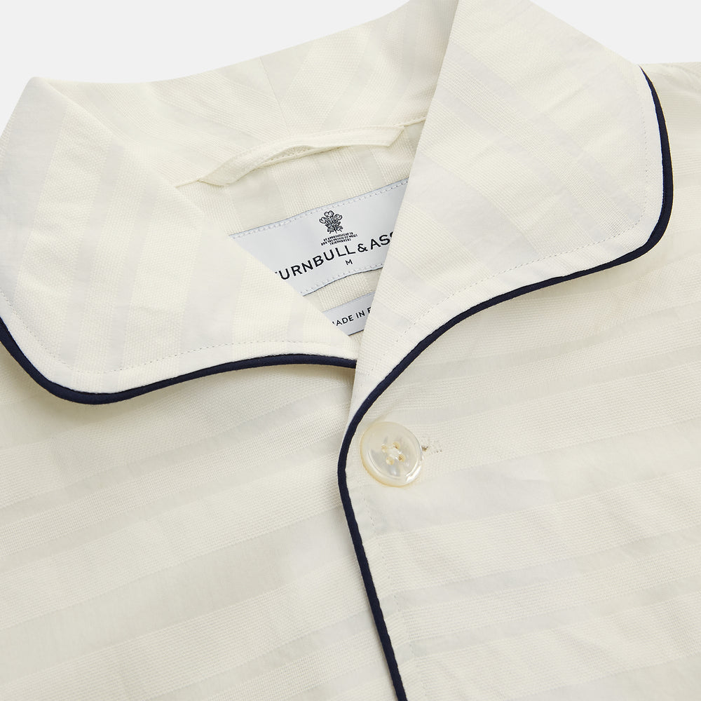 The Cream Pattern Cotton Blend Quin Pyjama Set by Turnbull & Asser features cream stripes, a long-sleeve button-up camp collar shirt with a chest pocket and black piping, and matching drawstring pants.