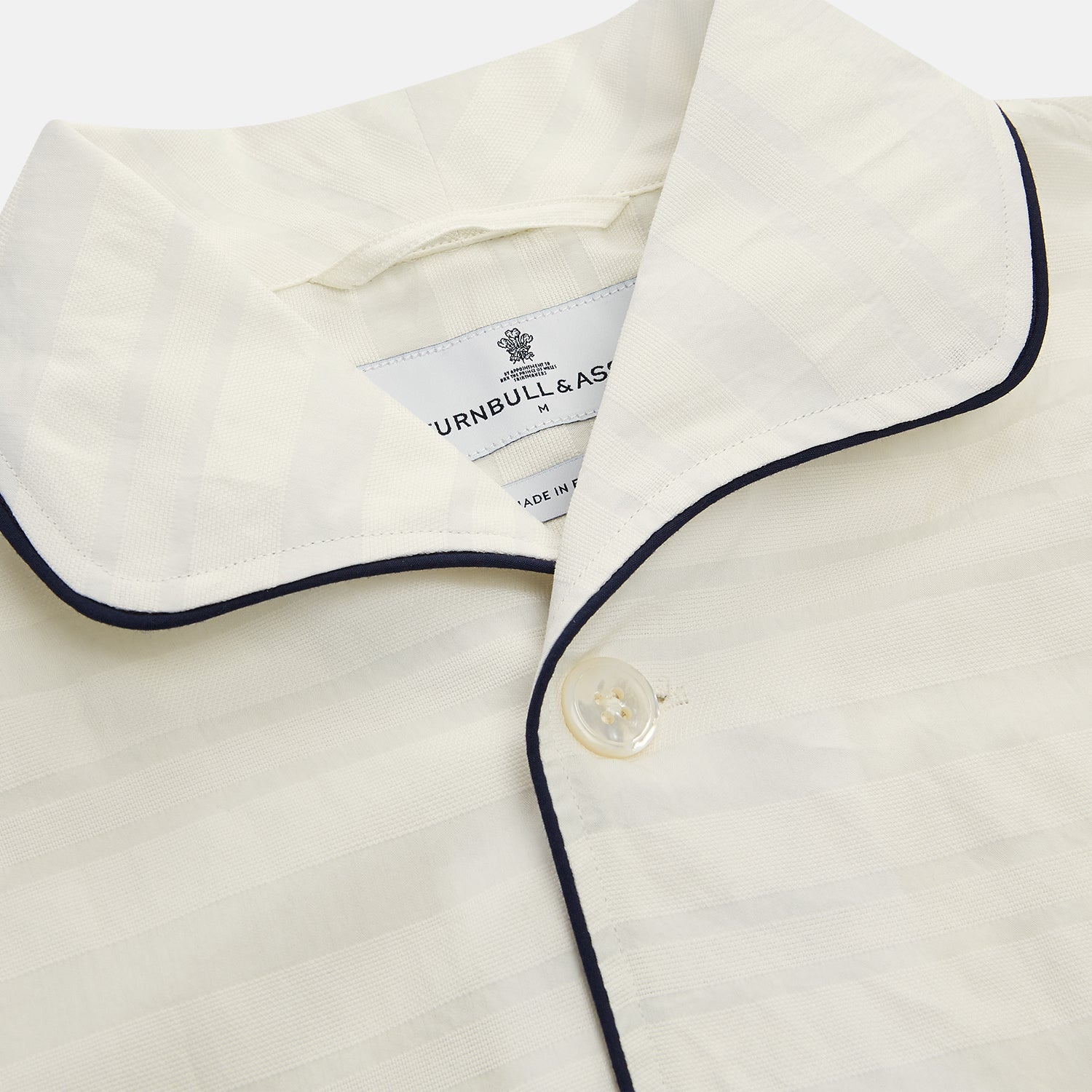 View 2 of product The Cream Pattern Cotton Blend Quin Pyjama Set by Turnbull & Asser features cream stripes, a long-sleeve button-up camp collar shirt with a chest pocket and black piping, and matching drawstring pants.