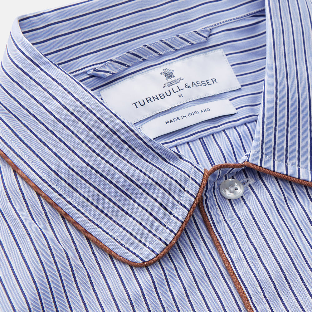 The Blue and Navy Halo Stripe Cotton Sussex Nightshirt by Turnbull & Asser features long sleeves, a collar, button-down front, chest pocket, curved hem, and brown piping for a classic luxury sleepwear style.