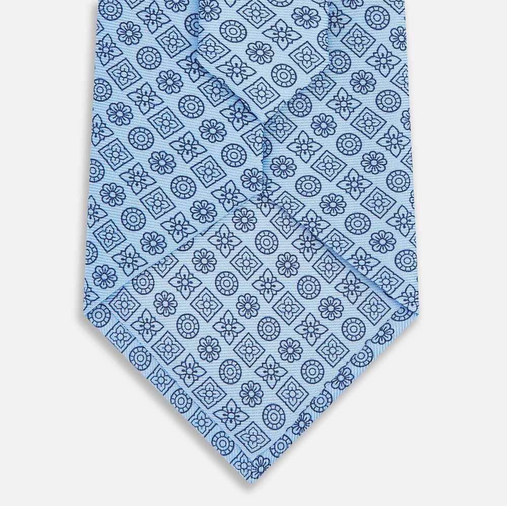The Turnbull & Asser Blue Floral Tile Pattern Printed Silk Tie features a light blue geometric and floral design in dark blue, shown rolled up on a white background.