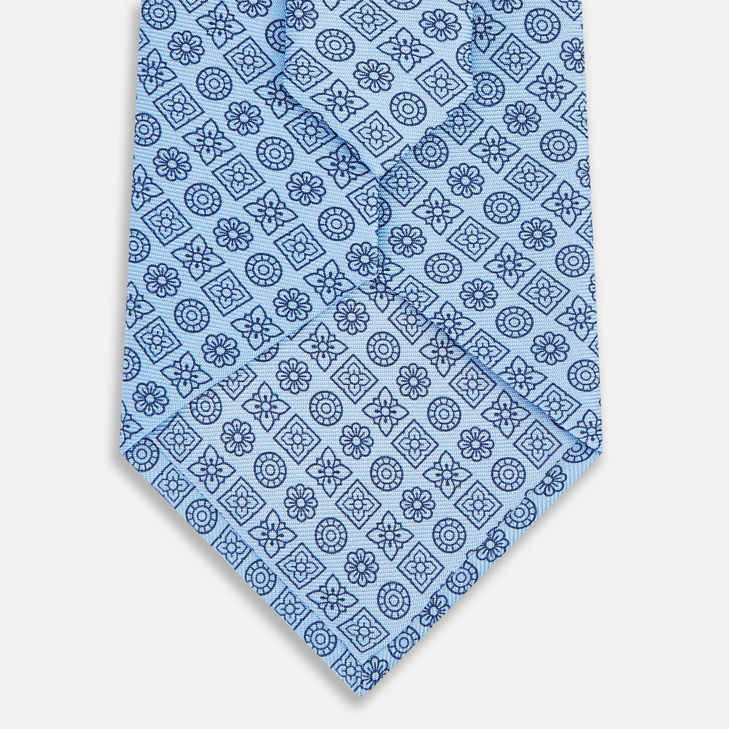 View 3 of product The Turnbull & Asser Blue Floral Tile Pattern Printed Silk Tie features a light blue geometric and floral design in dark blue, shown rolled up on a white background.