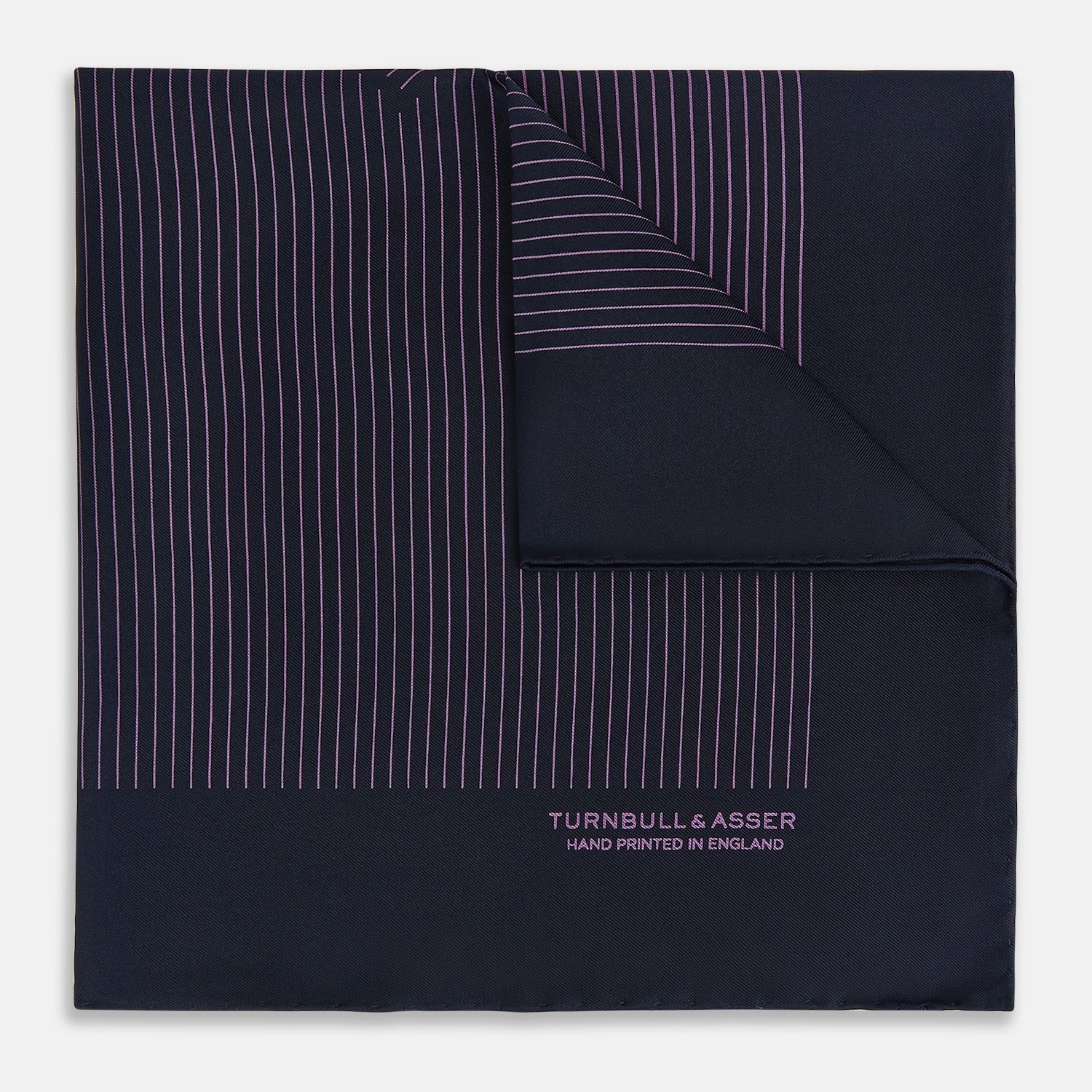 View 1 of product A navy silk pocket square with thin, even purple stripes and hand-rolled edges from Turnbull & Asser, featuring "Turnbull & Asser Hand Printed in England" printed in the corner.