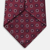 View 4 of product The Red Spheric Emblem Tie by Turnbull & Asser is a rolled-up maroon silk tie with a jacquard pattern of small white and purple circular dots, shown against a white background.